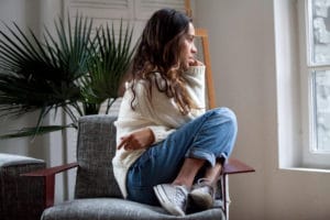 Mulher jovem sentada em poltrona olhando pela janela com expressão triste e pensativa ilustrando problema de saúde mental